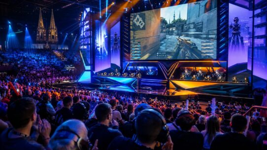 Large esports arena filled with spectators watching two teams compete on stage beneath a giant screen displaying a first-person shooter game