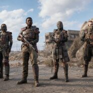 Four armored warriors in post-apocalyptic scrap-metal gear stand armed in a barren, rocky wasteland