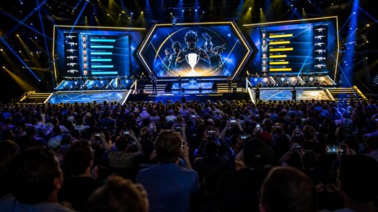 Large esports arena crowd facing a lit stage with giant screens displaying player stats and a trophy graphic during a Counter-Strike tournament