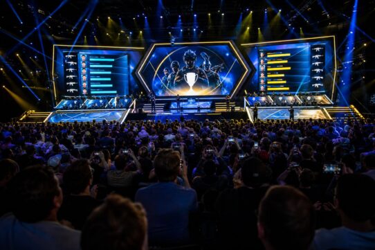 Large esports arena crowd facing a lit stage with giant screens displaying player stats and a trophy graphic during a Counter-Strike tournament